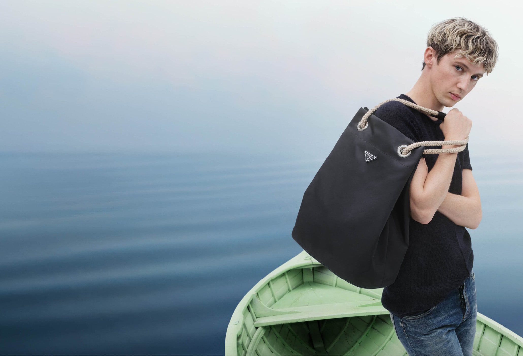 Troye Sivan standing on a green boat holding a black shoulder bag against an open misty water background.