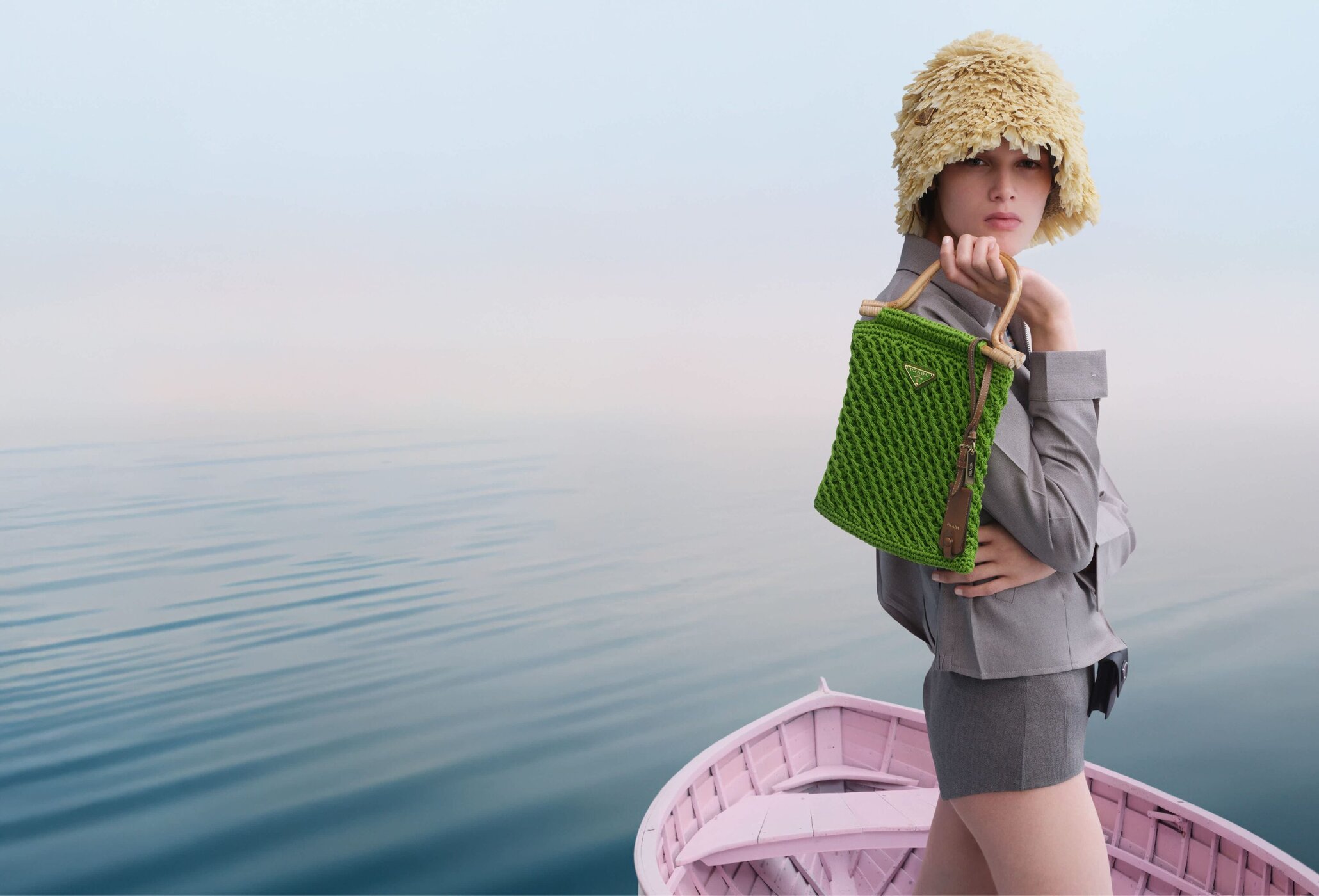 Model wearing textured yellow hat holding a green woven handbag on a pink boat against calm water.