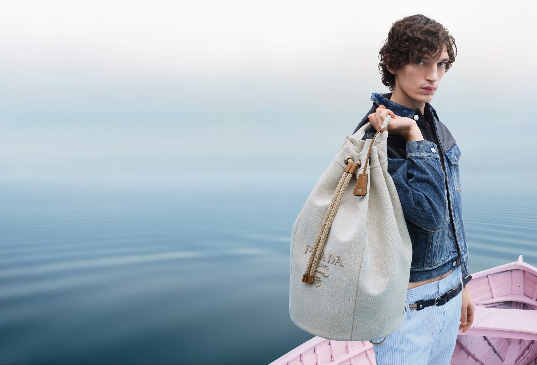 Model wearing denim jacket and pinstriped trousers holding large canvas bucket bag on pink boat over water