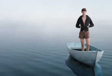 Young man standing inside a pale blue wooden rowboat on calm water under a misty overcast sky