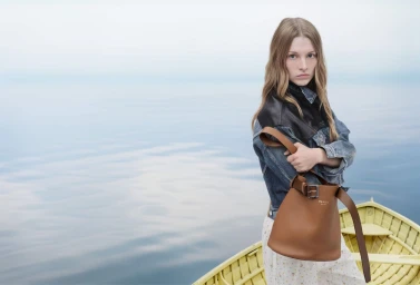 Model wearing a denim and leather jacket holding a brown leather bucket bag while standing in a yellow wooden boat on open water