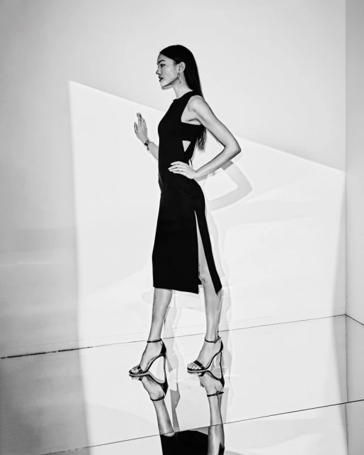 A slender, elegant woman in a black dress and high heels poses in a photo studio, with sleek hair and a modern, minimalist aesthetic.