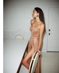 Model wearing a glittering rose gold spaghetti strap evening gown with a high slit standing beside a white dining table.