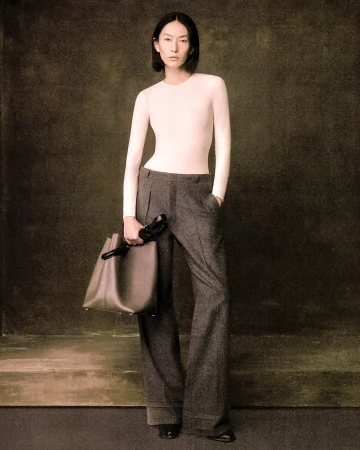 Model standing against a textured dark backdrop wearing a white long-sleeve top and grey trousers, holding a handbag.