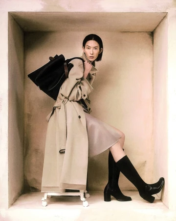 Model wearing a beige trench coat and black boots carrying a large black handbag inside a recessed wall alcove