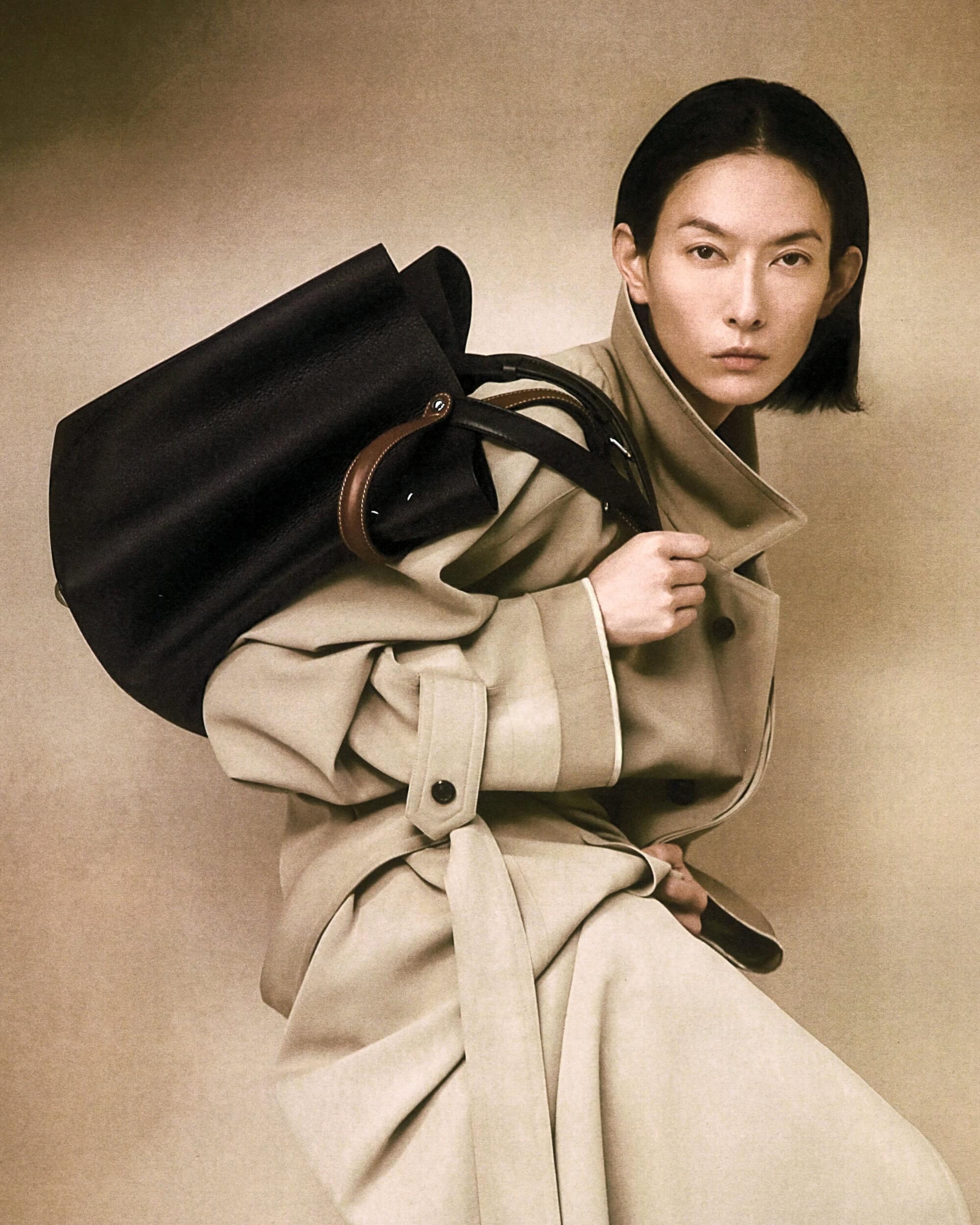 Asian woman model wearing a beige trench coat carrying a large dark handbag for Maison Margiela Spring 2025 campaign