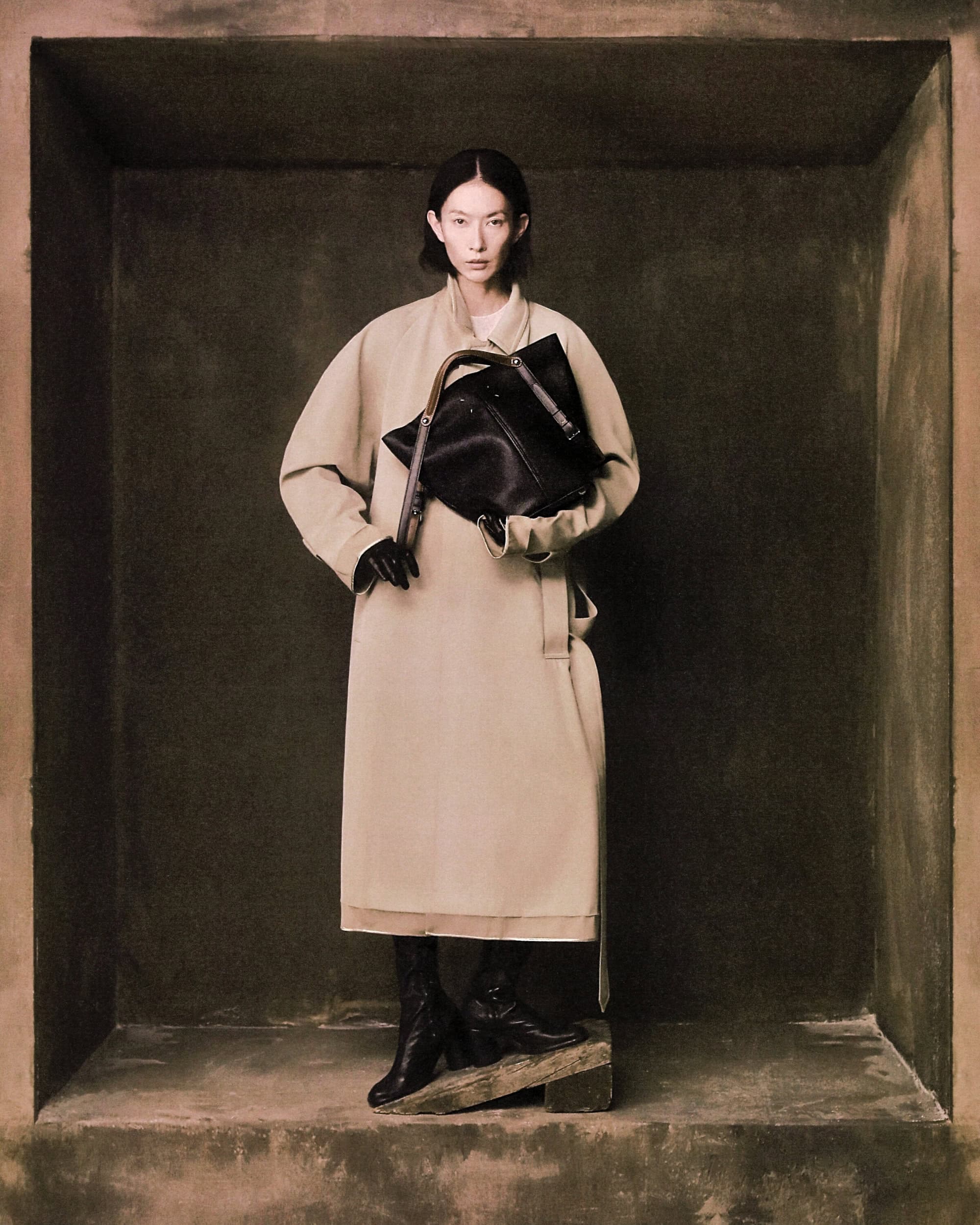 Model in long beige coat holding black handbag standing inside a dark textured architectural alcove