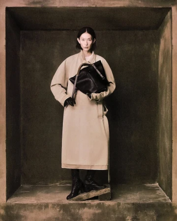 Model in long beige coat holding black handbag standing inside a dark textured architectural alcove