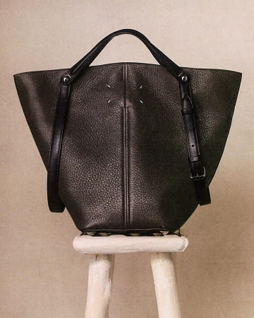 Maison Margiela black leather tote bag resting on a weathered white wooden stool against a neutral wall.