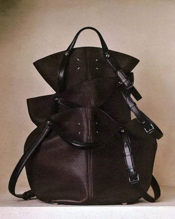 Three dark brown leather Maison Margiela handbags stacked vertically on a pale neutral studio surface