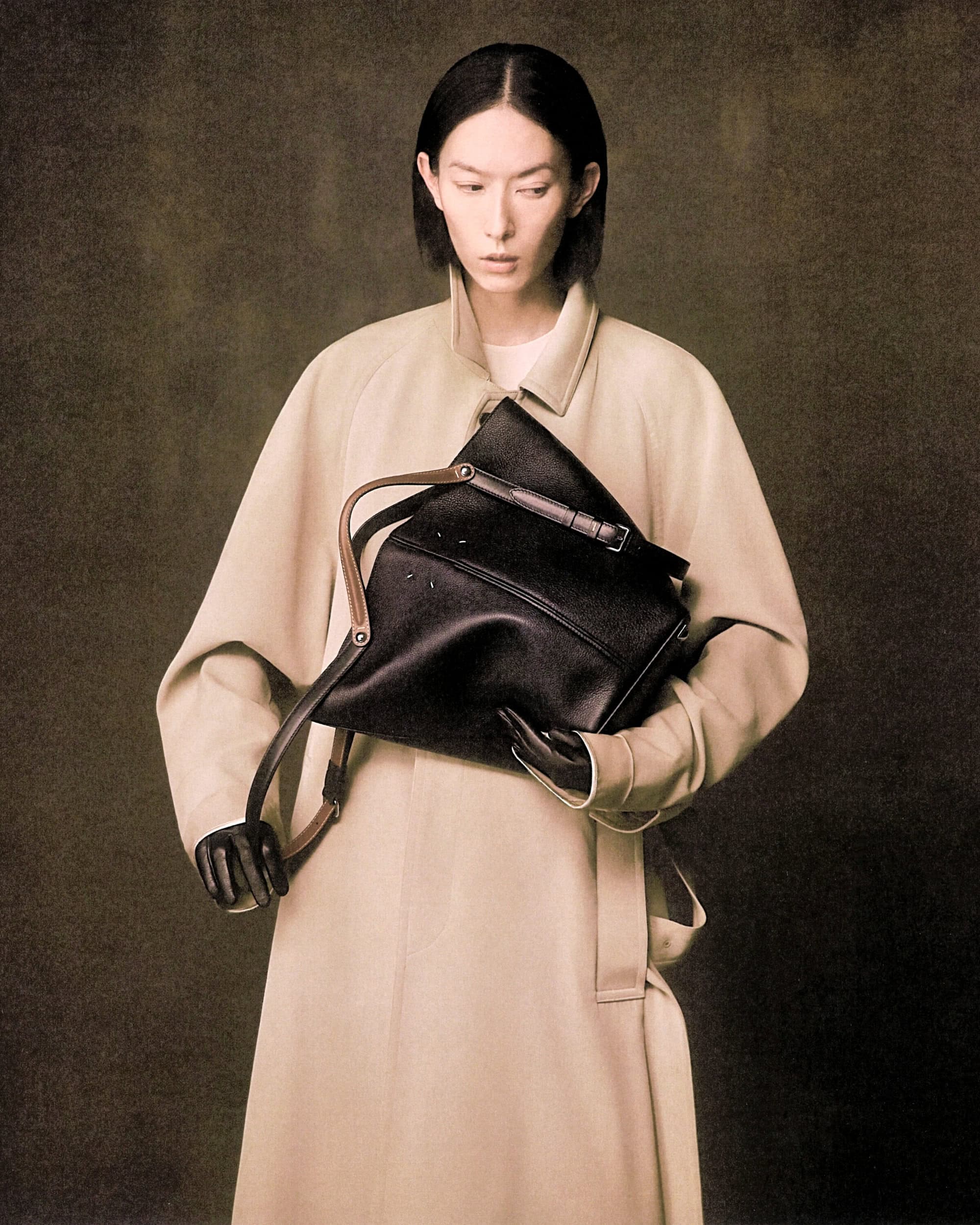 Model in a tan coat holding a black leather shoulder bag against a textured backdrop for Maison Margiela.