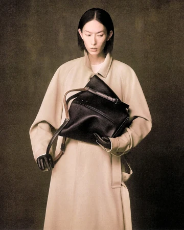 Model in a tan coat holding a black leather shoulder bag against a textured backdrop for Maison Margiela.