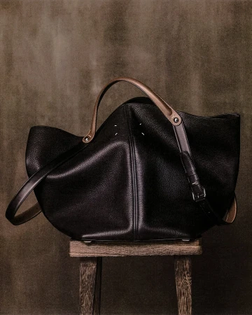 Black leather designer handbag resting on a small wooden stool against a textured olive background.