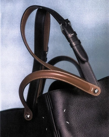 Close up of Maison Margiela leather tote bag with contrasting brown handles and black strap against textured backdrop