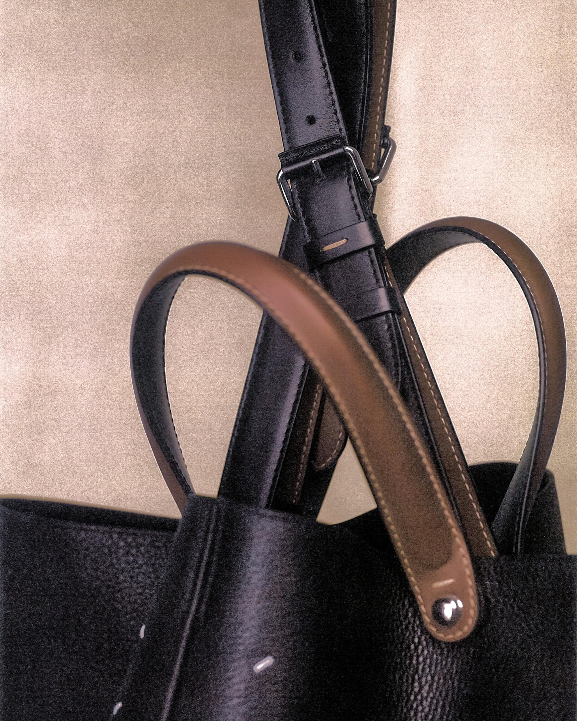 Close-up of Maison Margiela black leather handbag with brown handles and adjustable strap against textured backdrop.