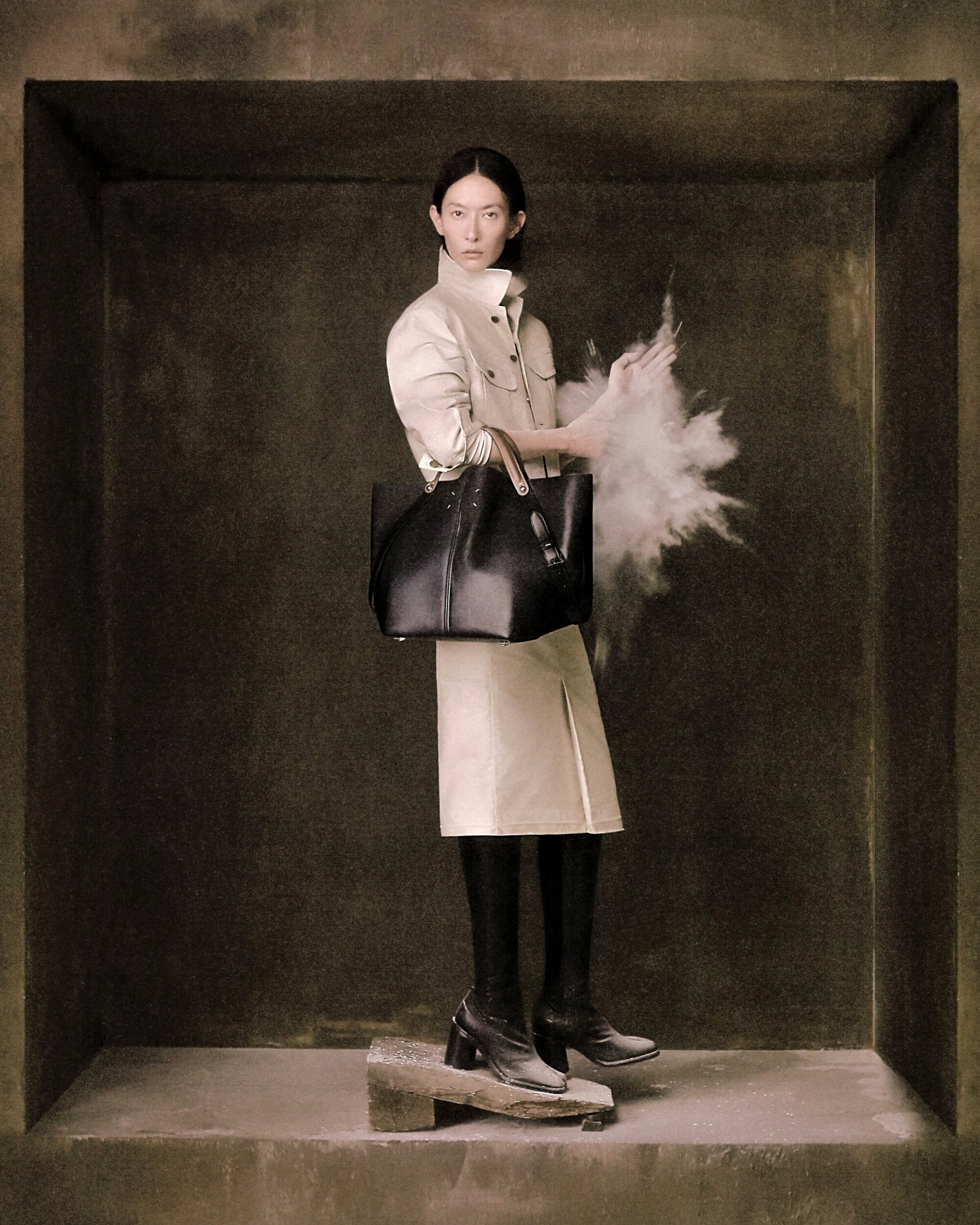 Model standing inside a stone alcove wearing a cream coat and black boots while holding a large dark tote bag.
