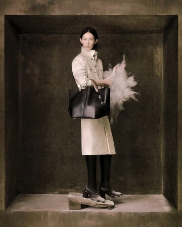 Model standing inside a stone alcove wearing a cream coat and black boots while holding a large dark tote bag.