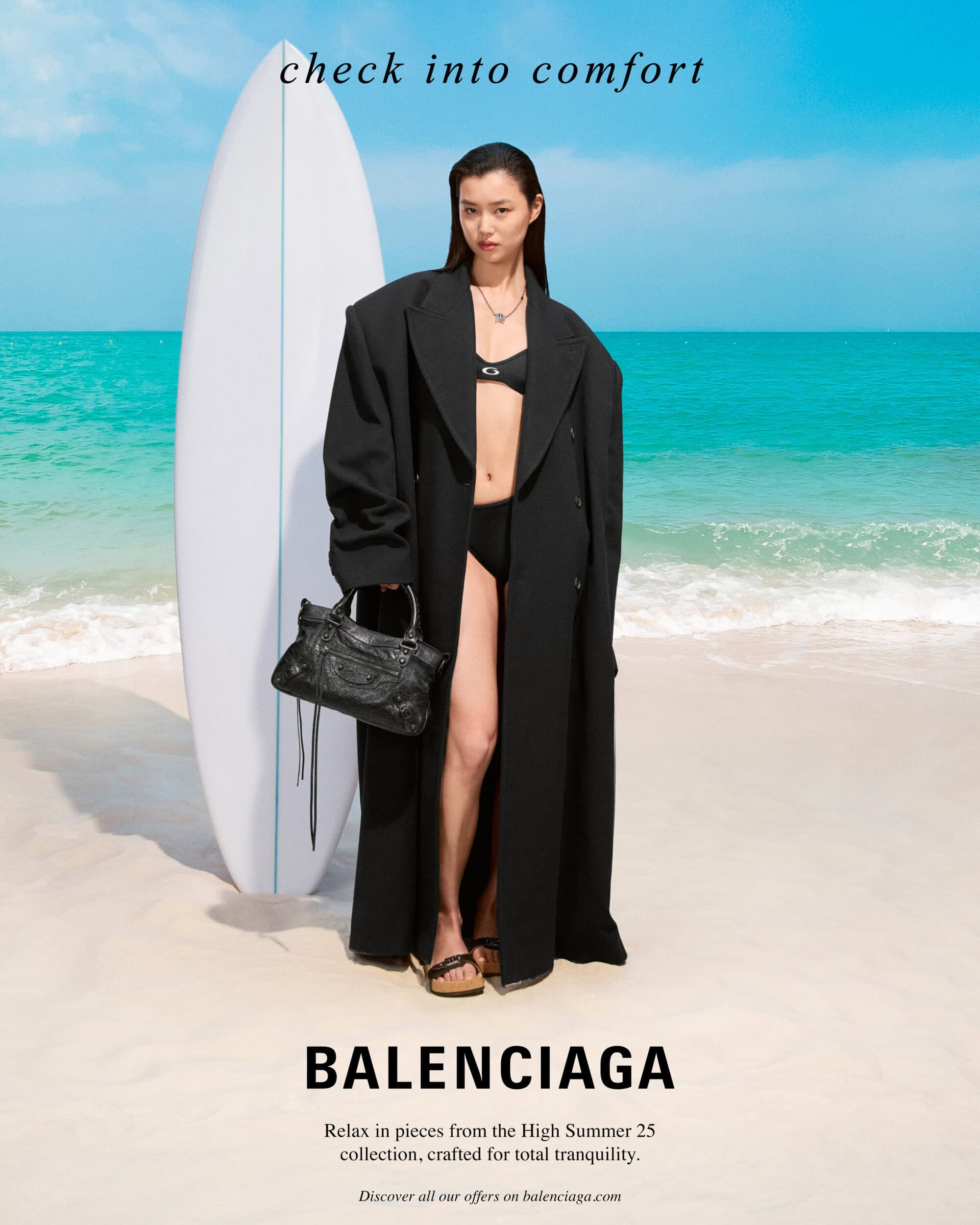 Model standing on a sandy beach wearing a black coat over a bikini, holding a black handbag next to a surfboard.
