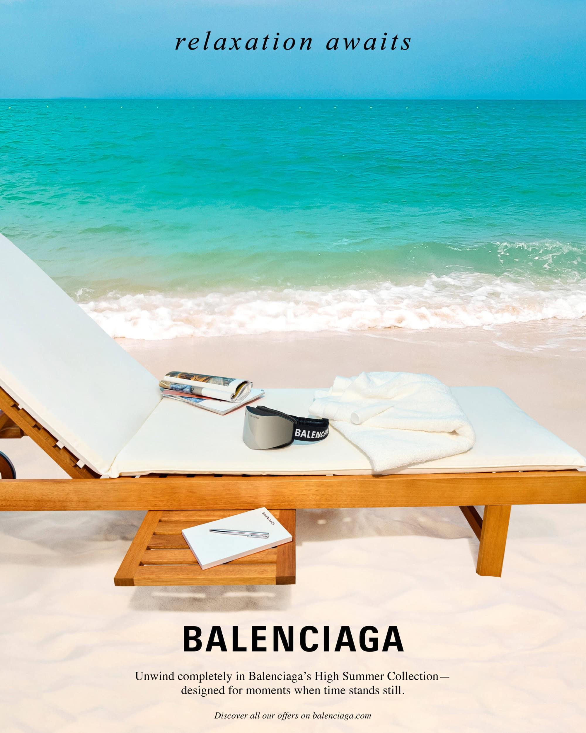 Wooden sun lounger on a sandy beach with sunglasses, magazines and a folded white towel against a turquoise ocean.