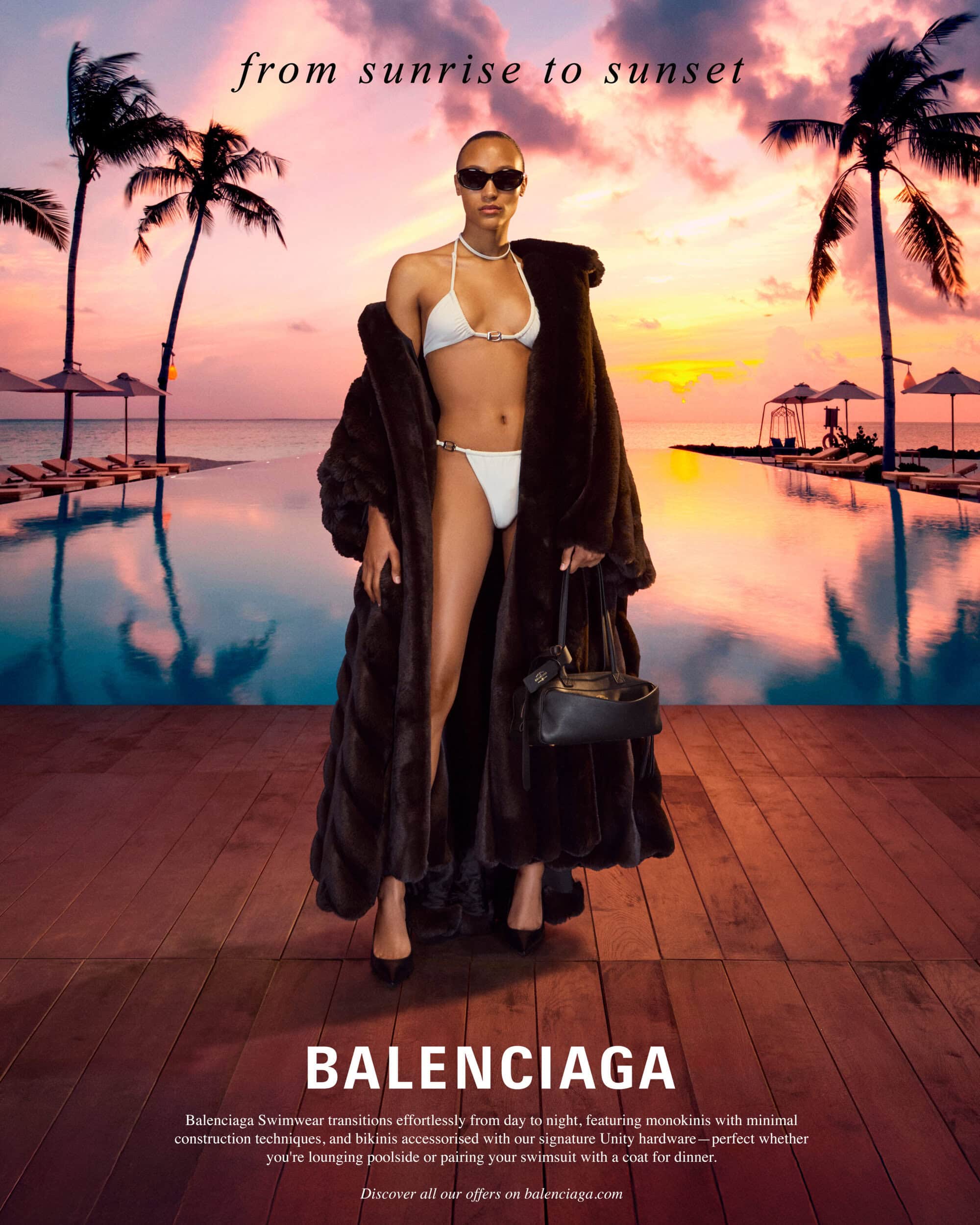 Model in white bikini and heavy dark coat standing by a pool at sunset holding a black handbag