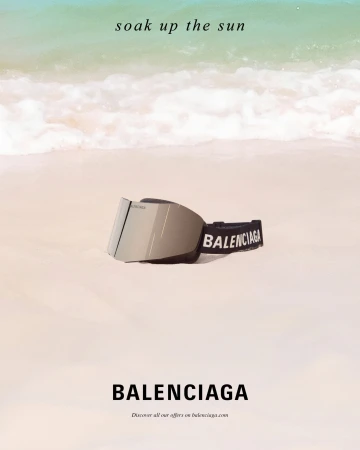 Balenciaga mirrored ski goggles resting on a light sandy beach with gentle ocean waves in the background.