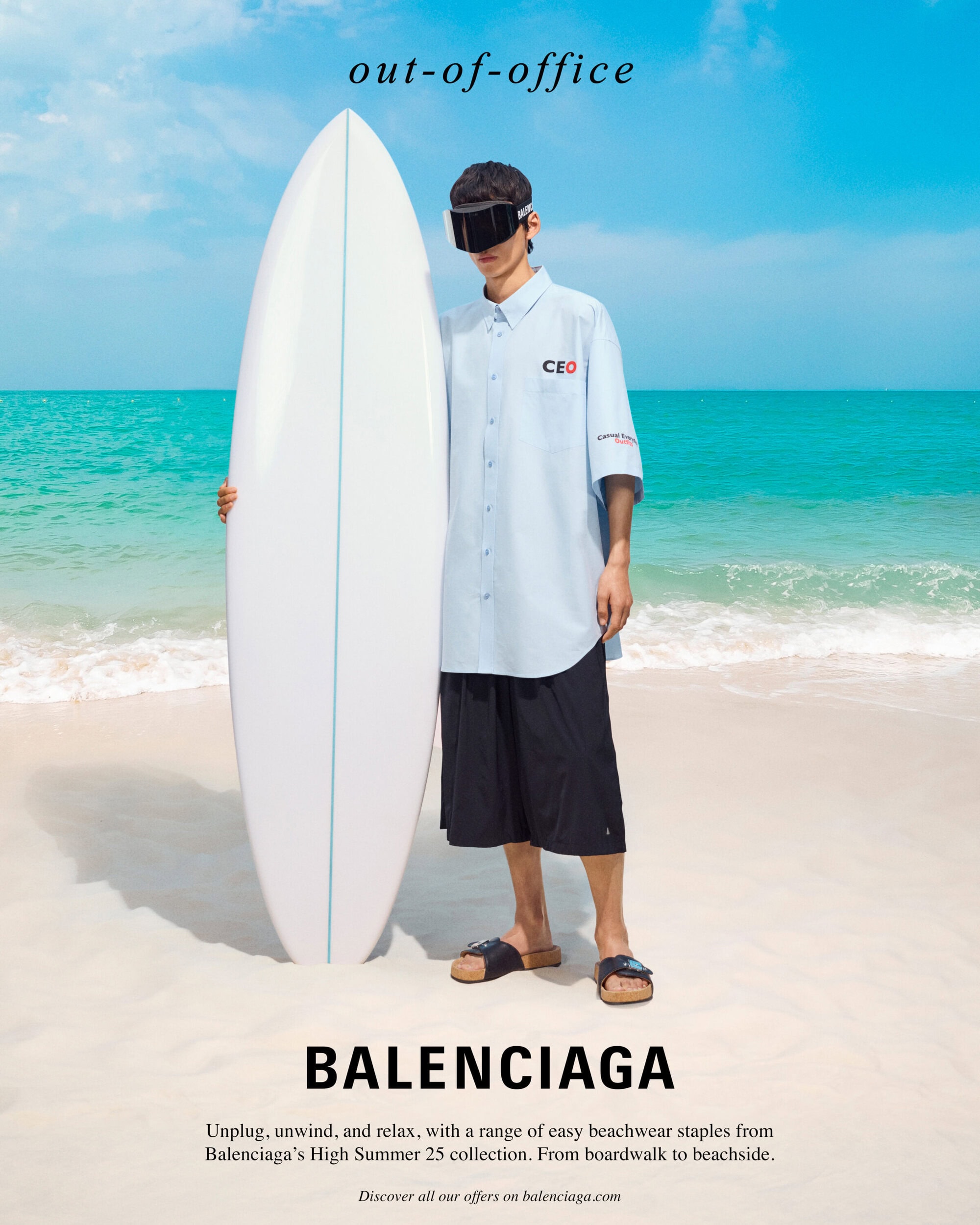 Man holding a white surfboard on a sandy beach wearing a light blue button-down shirt and black shorts