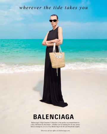 Model in a black maxi dress and sunglasses holding a straw tote bag on a sandy beach