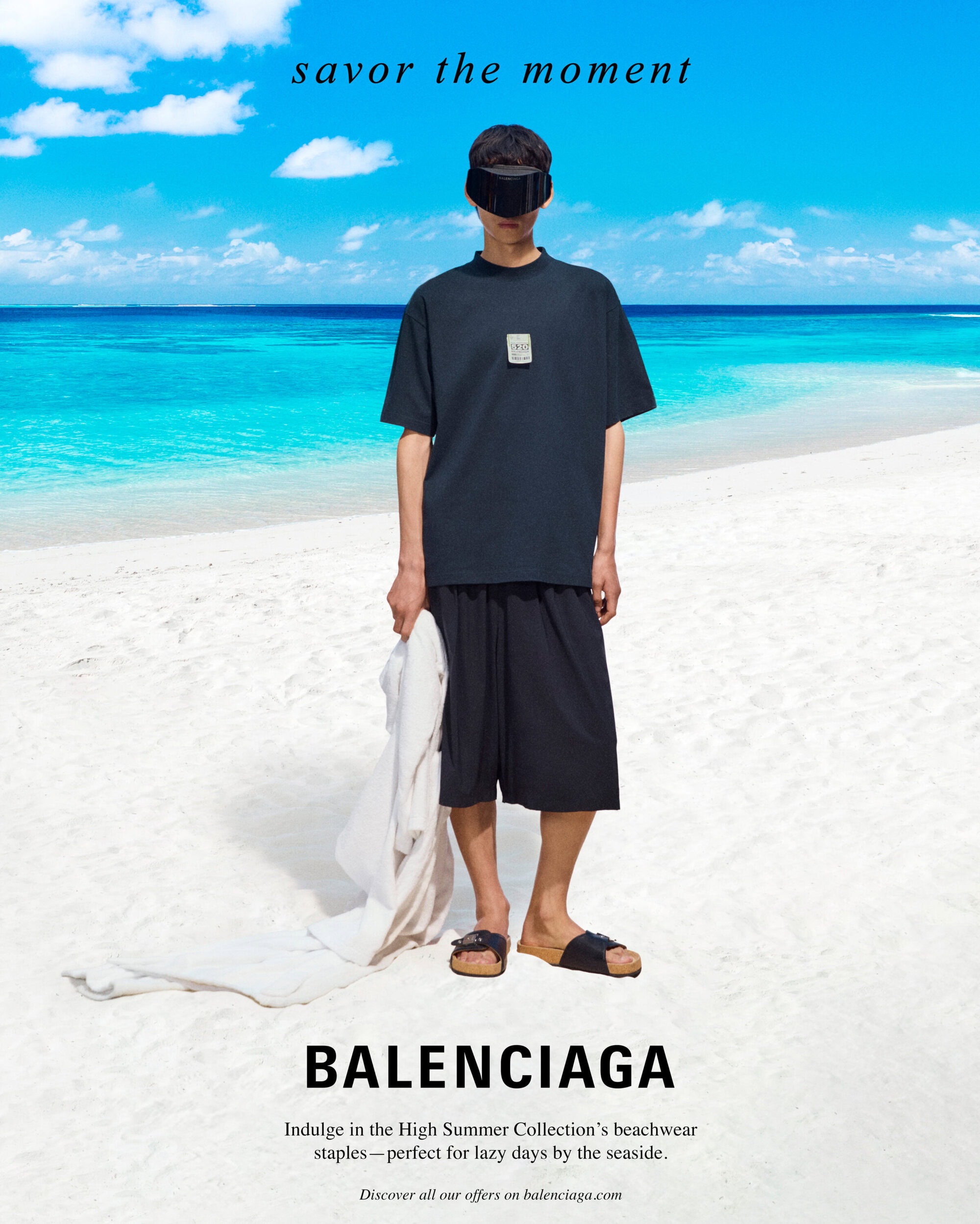 Man wearing a dark t-shirt, shorts, and eye covering standing on a white sand beach with turquoise ocean water in the background.