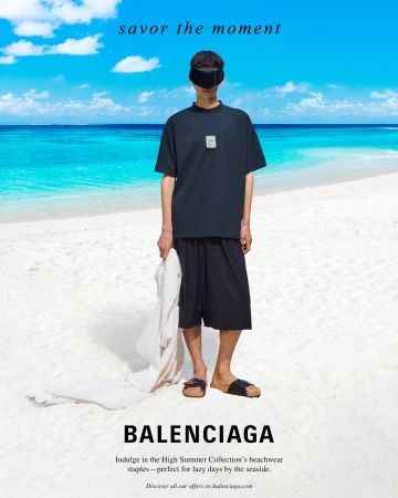 Man wearing a dark t-shirt, shorts, and eye covering standing on a white sand beach with turquoise ocean water in the background.