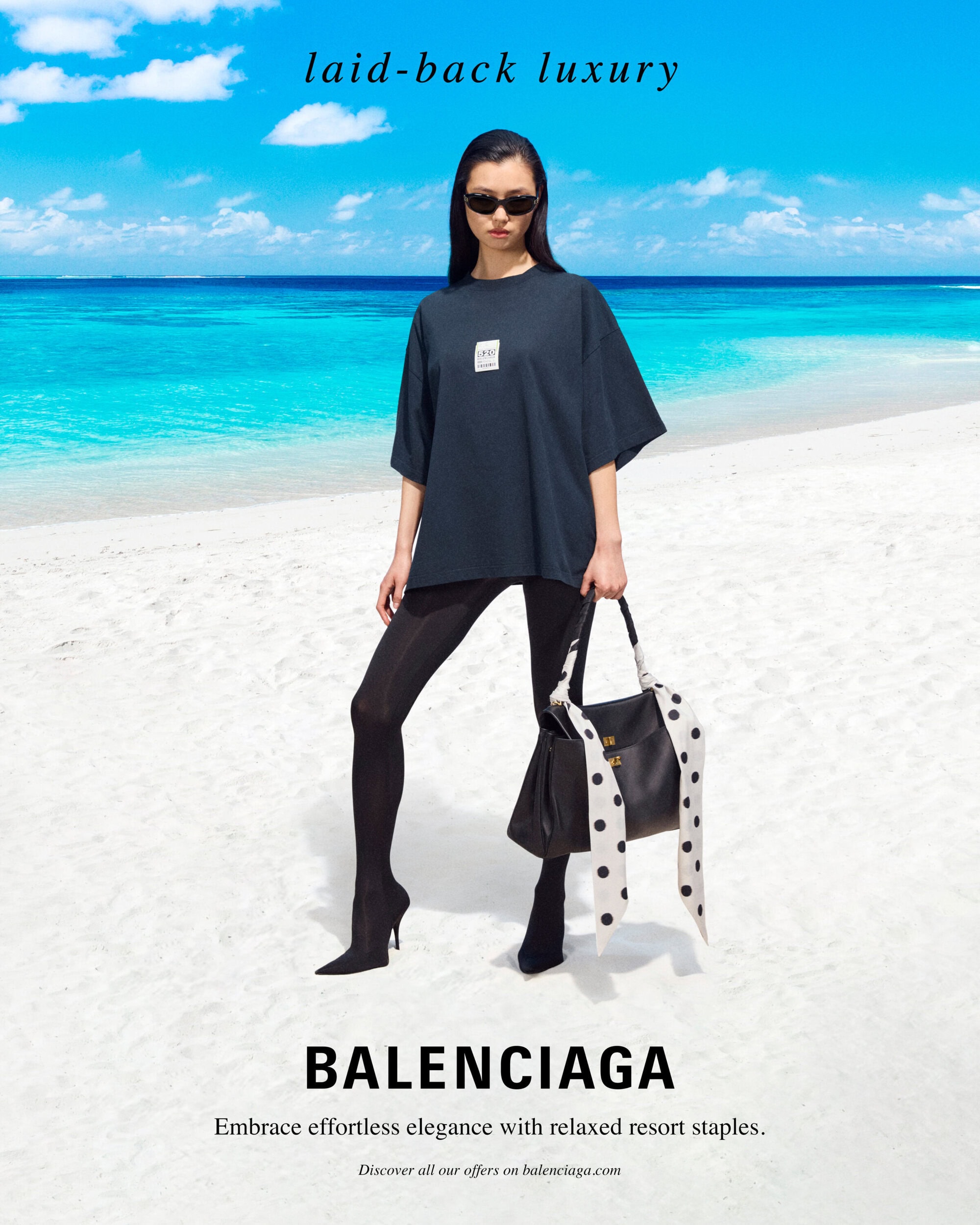 Model standing on a sandy beach wearing a dark t-shirt and holding a black handbag with polka-dot ribbons