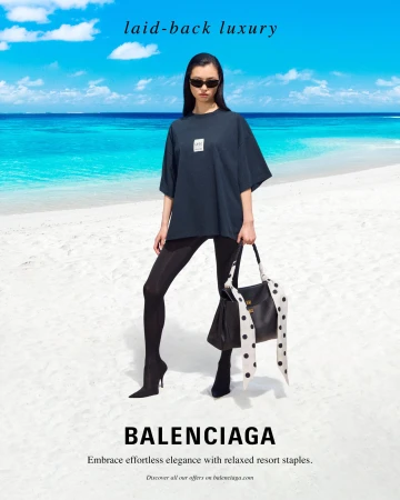 Model standing on a sandy beach wearing a dark t-shirt and holding a black handbag with polka-dot ribbons