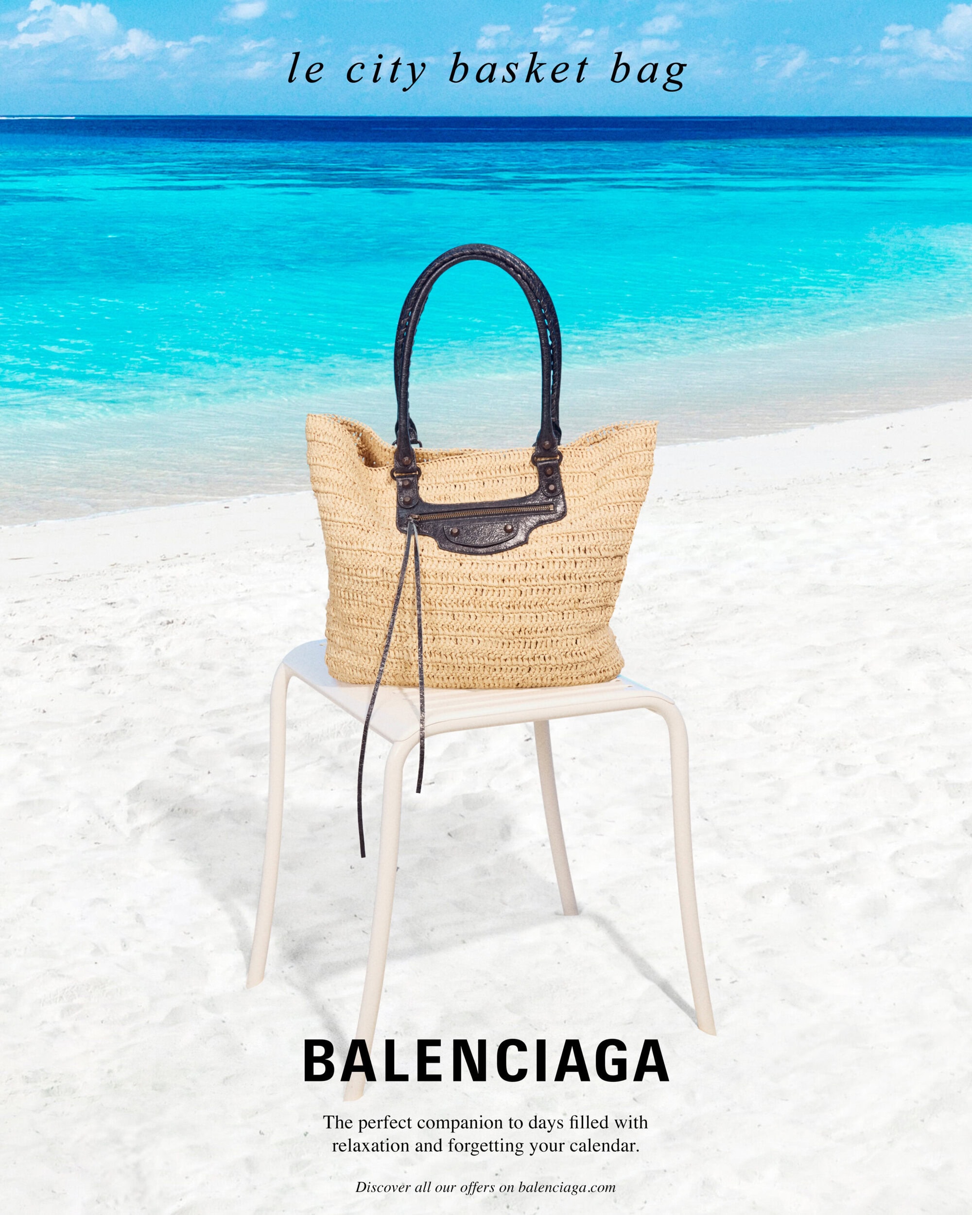 Straw basket bag with dark leather handles resting on a white chair on a sandy beach with bright blue ocean water