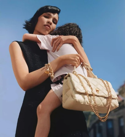 Model holding a child while carrying a quilted handbag against a bright clear sky