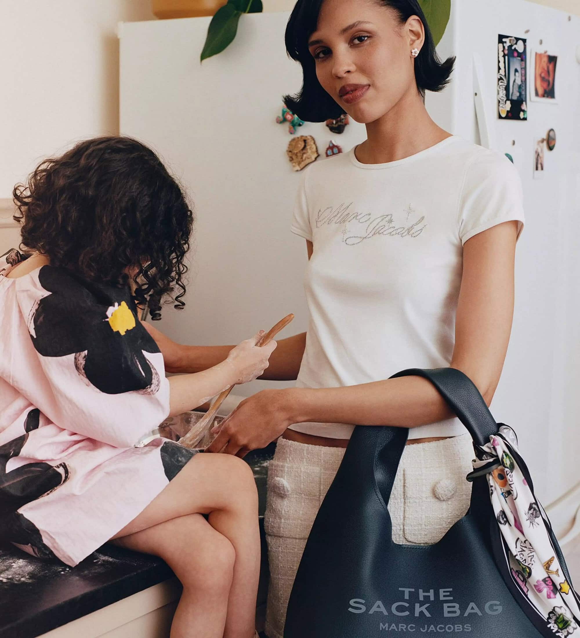 Model Nara Smith holding a black leather bag beside a child in a kitchen for Marc Jacobs Mother's Day campaign
