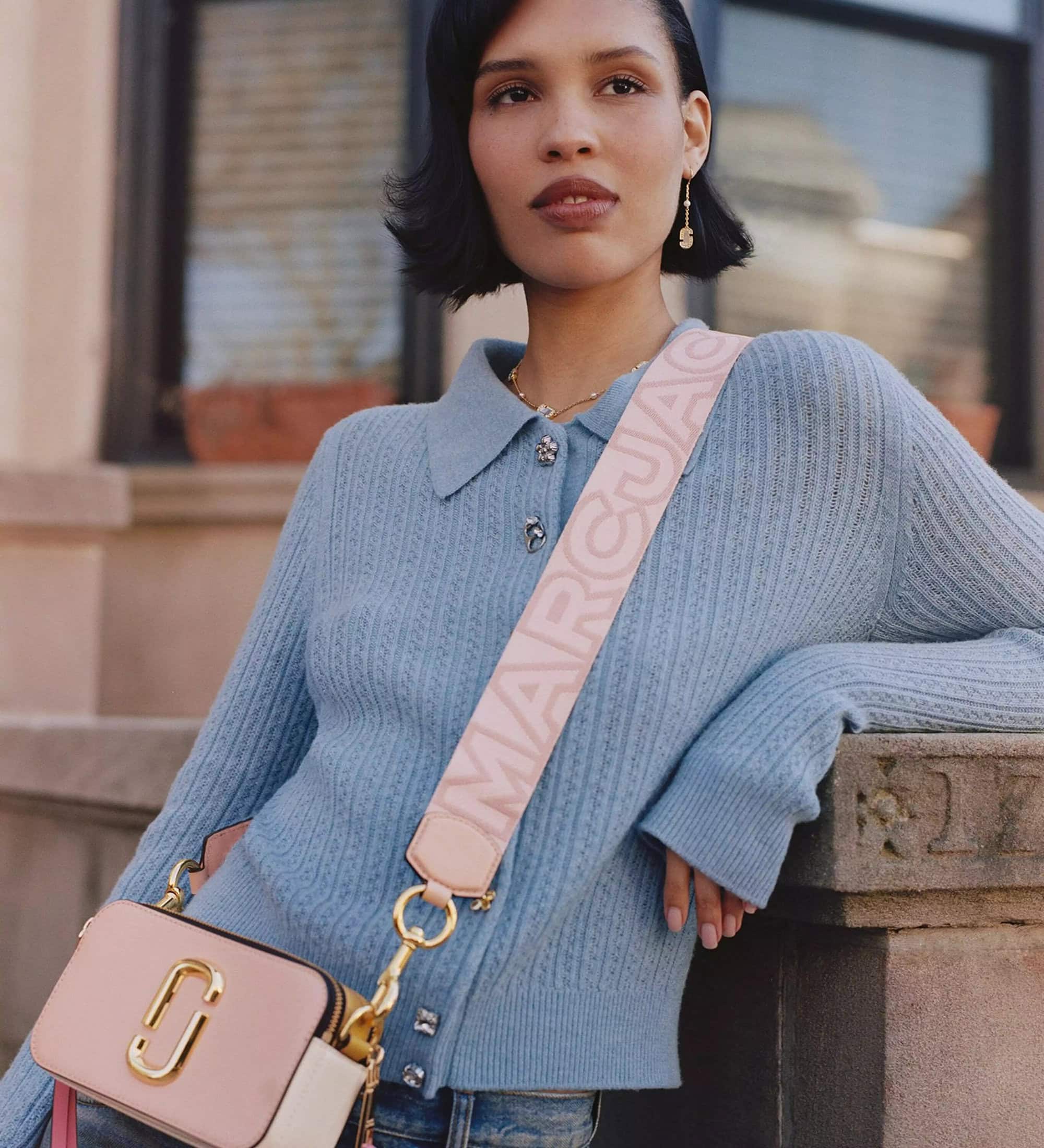 Model wearing a blue cardigan and pink branded crossbody bag leaning against a stone building exterior