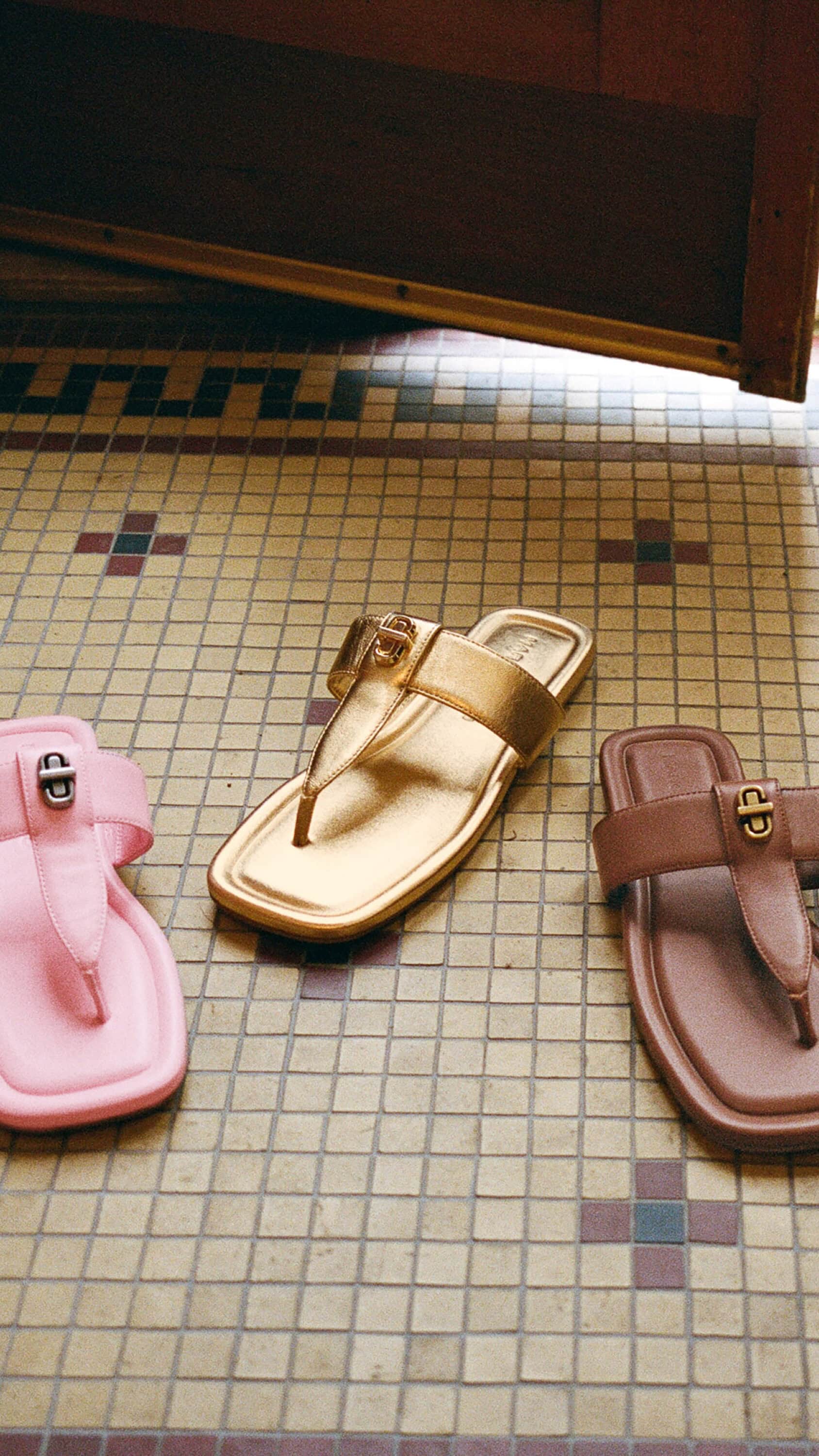 Three flat sandals in pink, gold, and brown resting on a tiled floor with geometric patterns.
