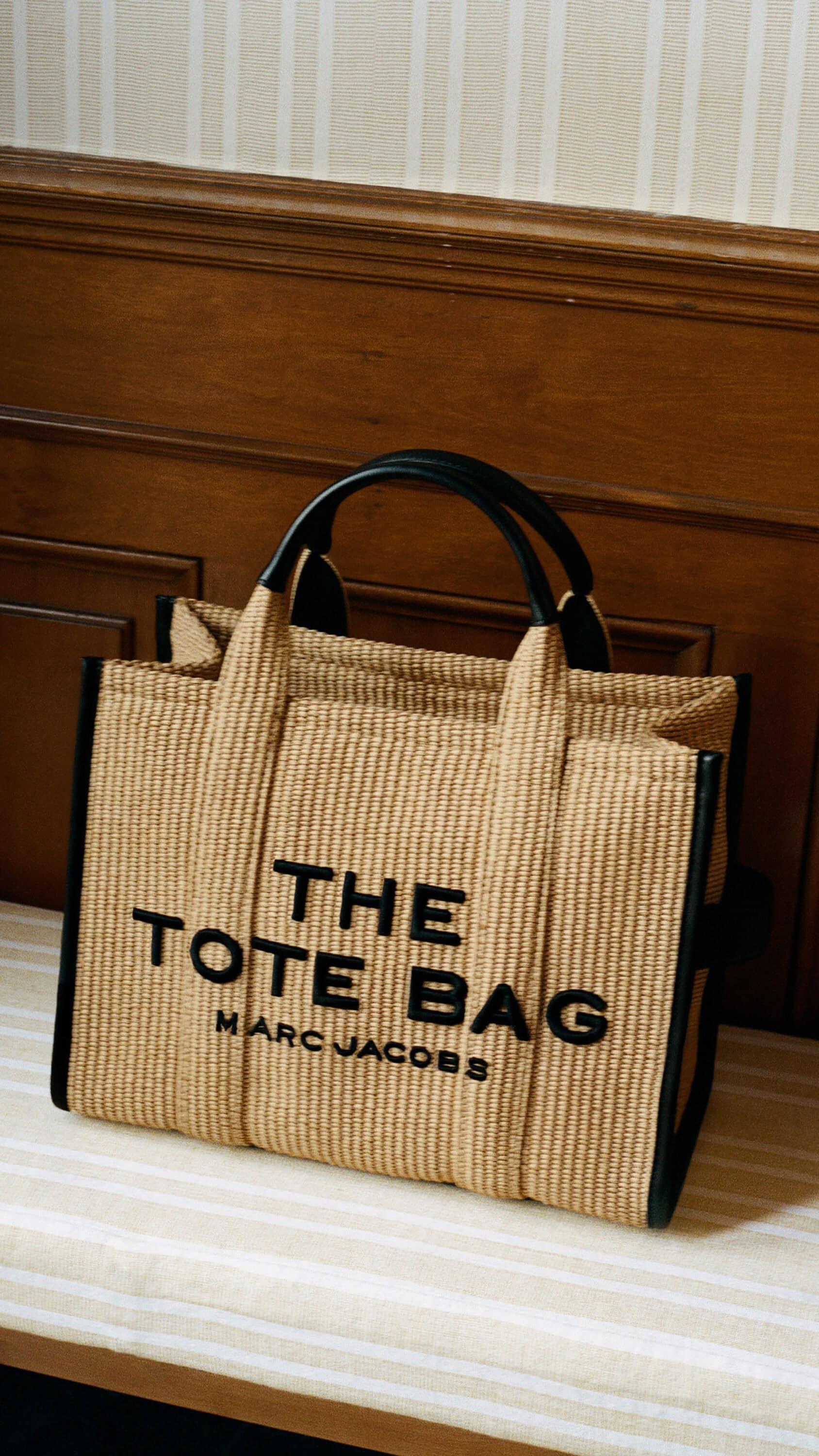 Marc Jacobs woven tote bag with black leather handles resting on a striped bench against wooden wainscoting.