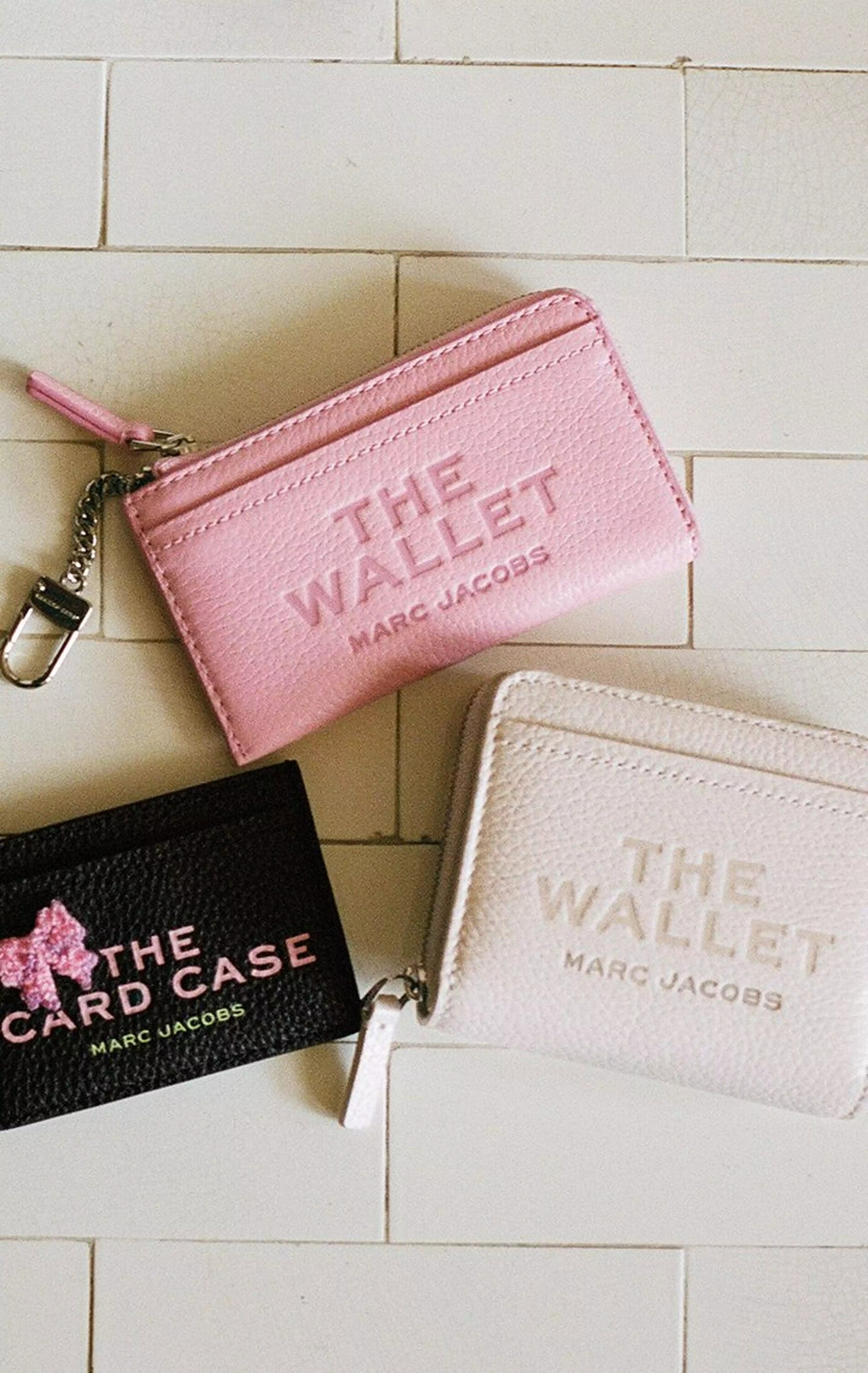 Three Marc Jacobs wallets placed on a light tiled surface, including a pink wallet, a white wallet, and a black card case.