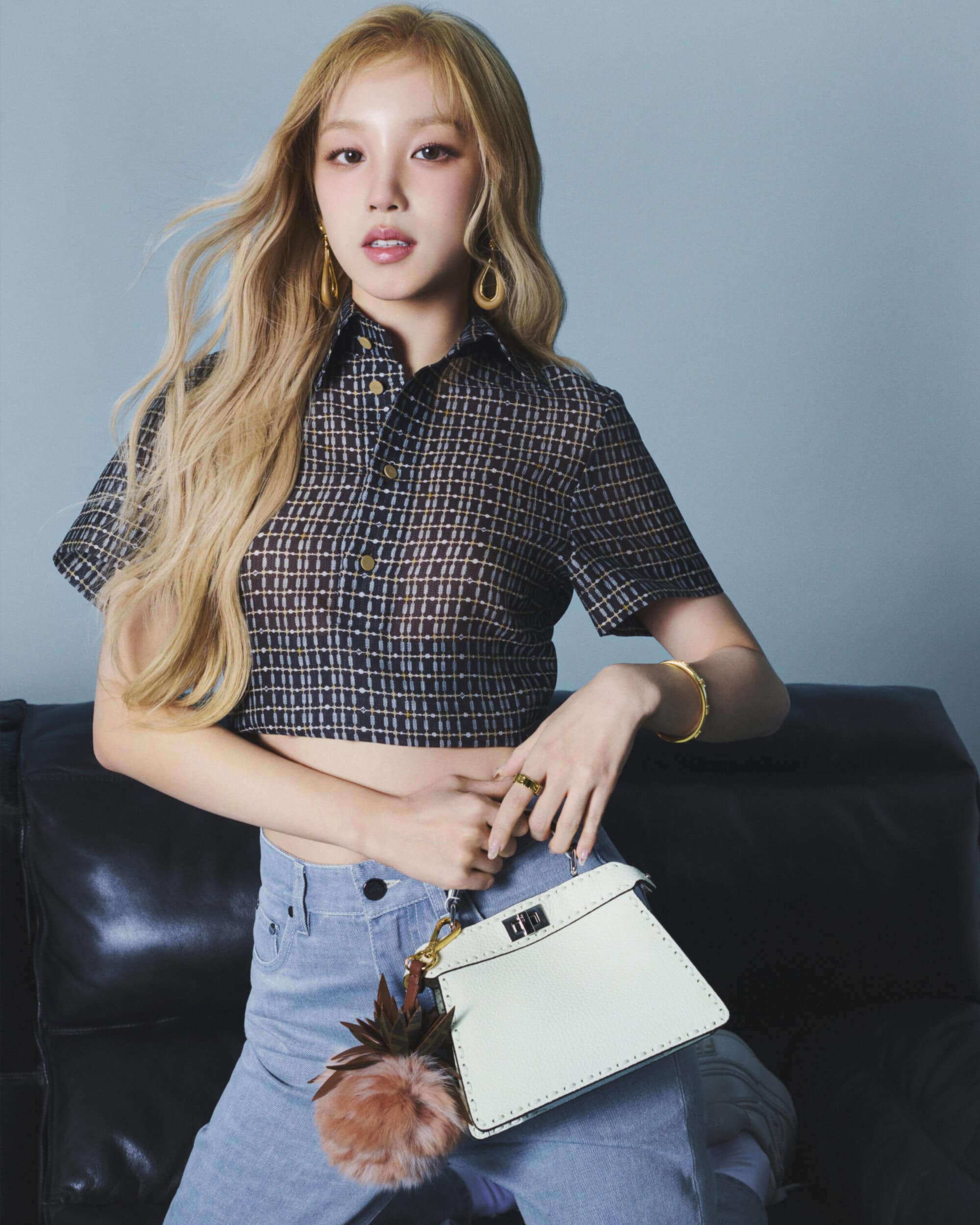 Song Yuqi holding a white leather handbag wearing a dark patterned short-sleeve shirt and light blue jeans against a grey wall.