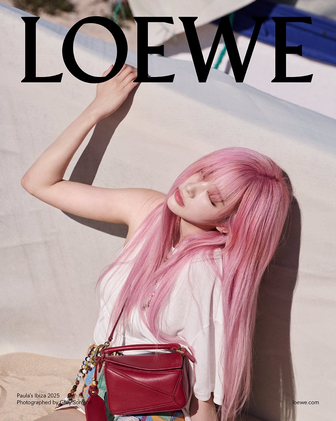 Model with long pink hair wearing a white top and holding a red handbag against a light background