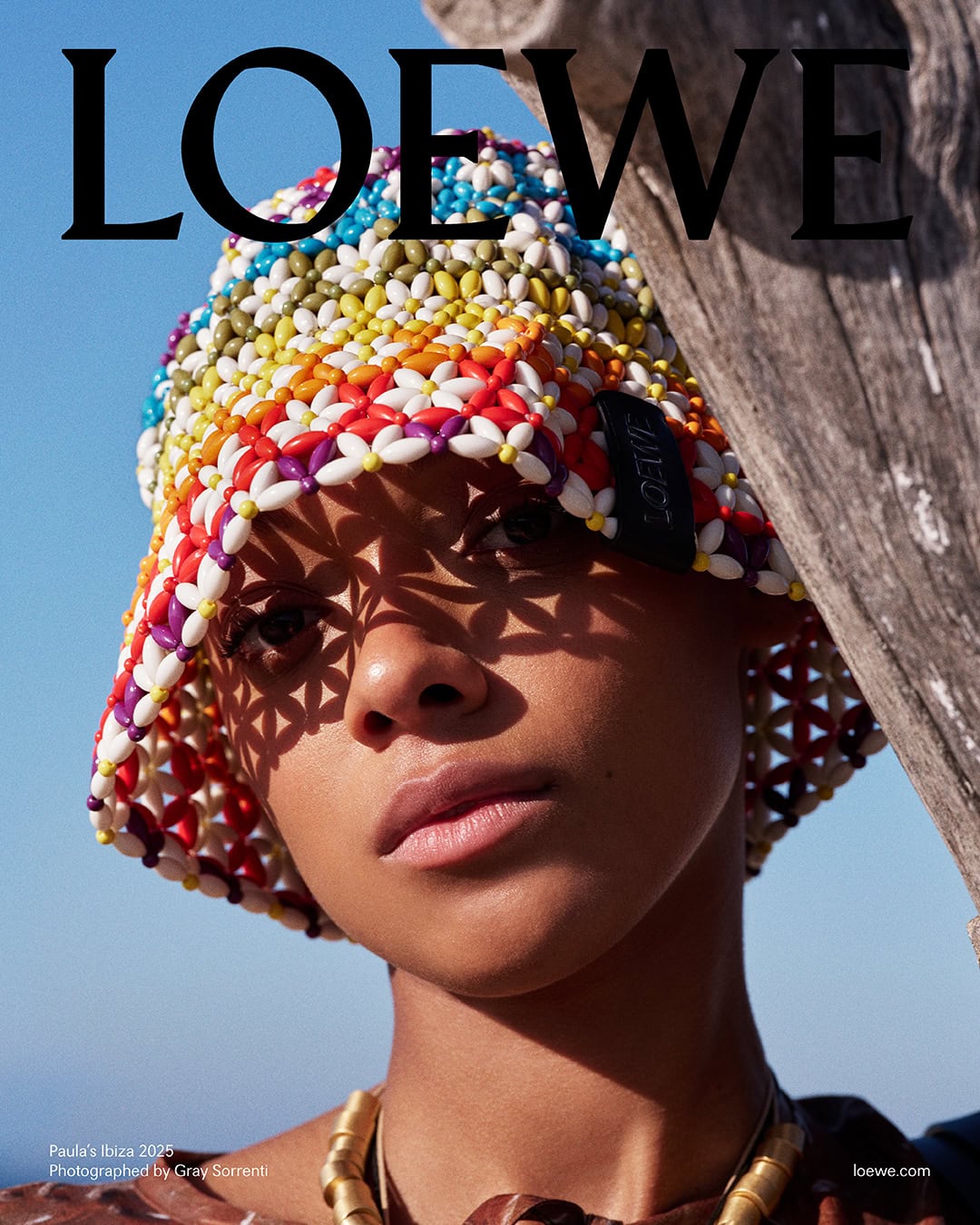 Model wearing a colourful beaded bucket hat with geometric flower patterns under bright outdoor sunlight against a clear blue sky