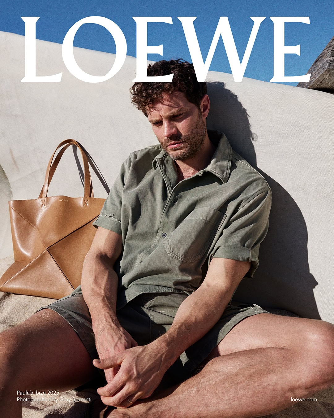 Jamie Dornan seated outdoors against a light wall wearing an olive green short-sleeve shirt with a large tan tote bag nearby.