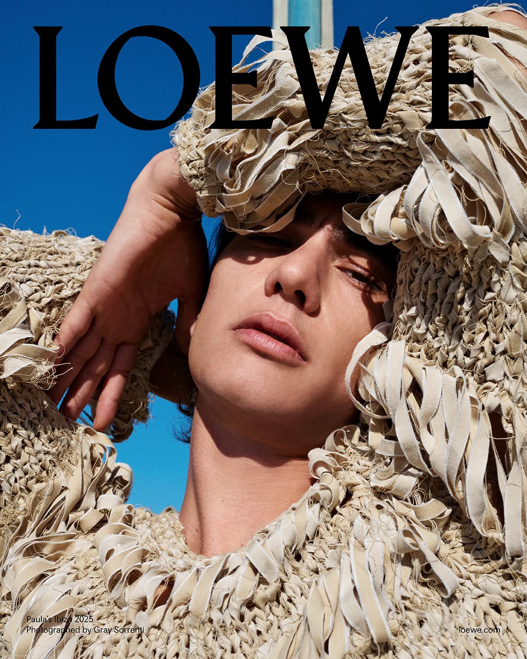 Close-up portrait of a model wearing a textured fringe garment against a clear blue sky