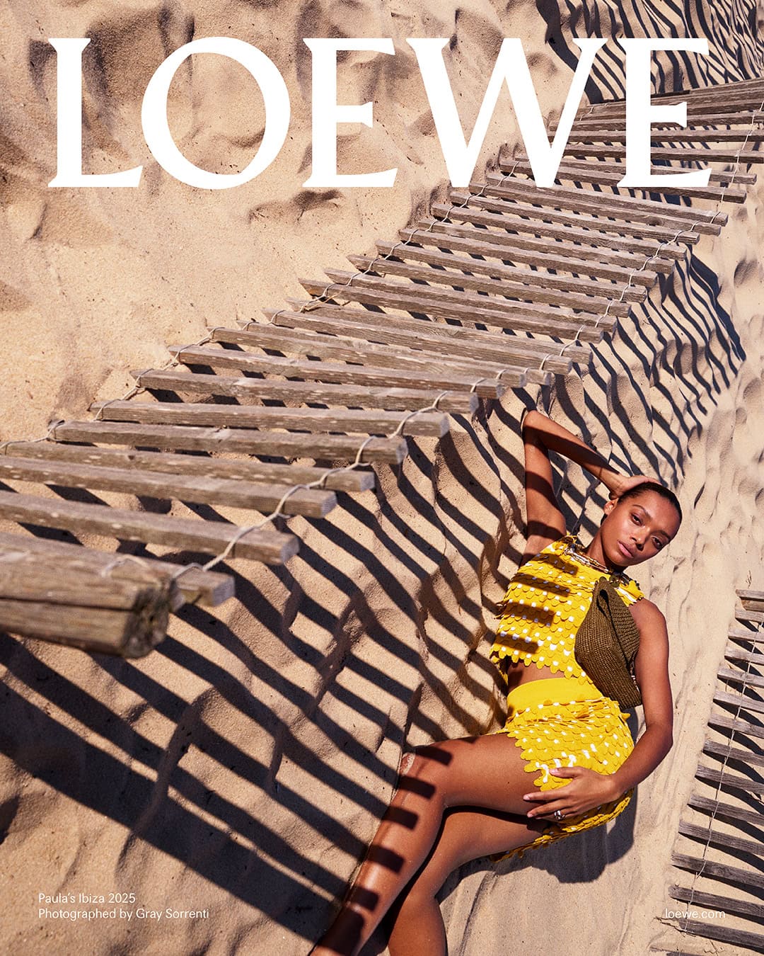 Model wearing yellow textured top and skirt lying on sandy beach beneath wooden boardwalk slats casting shadows.