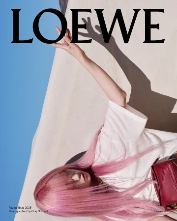 Model with long pink hair wearing a white top and holding a red handbag against a bright sky background.