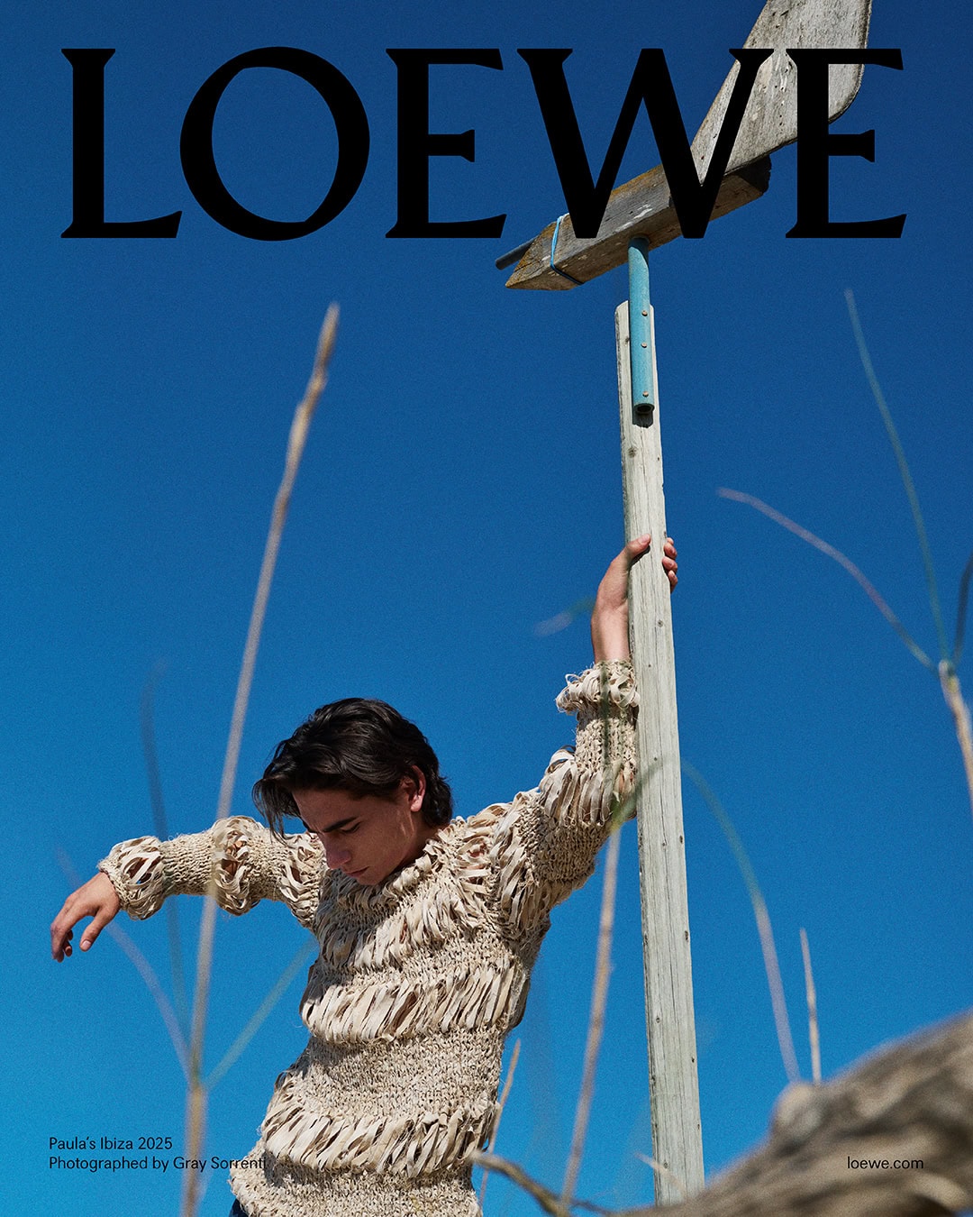 Man wearing a textured knit sweater holding onto a wooden pole against a clear blue sky for Loewe campaign