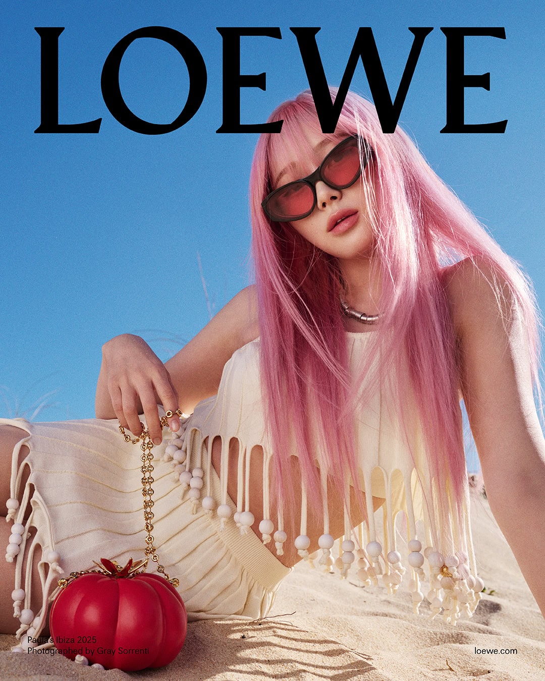 Woman with long pink hair wearing sunglasses and cream knit clothing holding a red tomato handbag on a sandy beach.