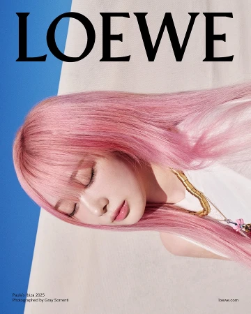 Close-up portrait of a woman with long pink hair wearing a white top and gold necklace against a clear blue sky.