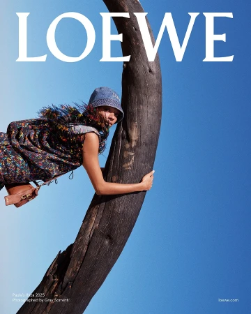 Model in patterned fringe top and bucket hat hugging a dark curved tree trunk against a clear blue sky