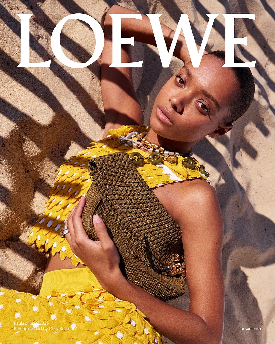 Model in yellow textured top holding a brown woven clutch resting on sandy ground